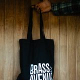 Ruka drží černou plátěnou tašku s logem Brass Avenue před dřevěnou stěnou