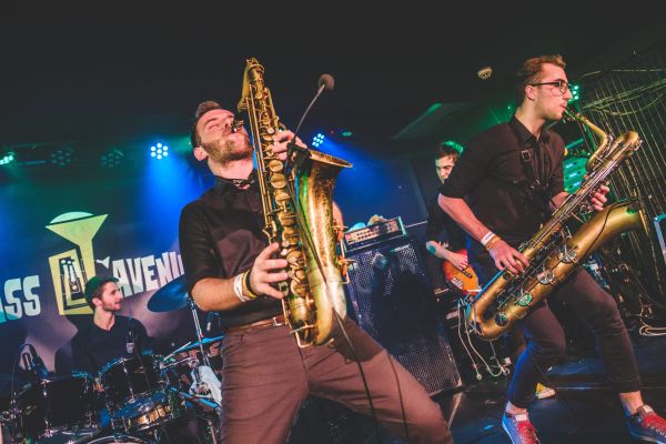 Dva saxofonisté na pódiu před bubeníkem a logem Brass Avenue